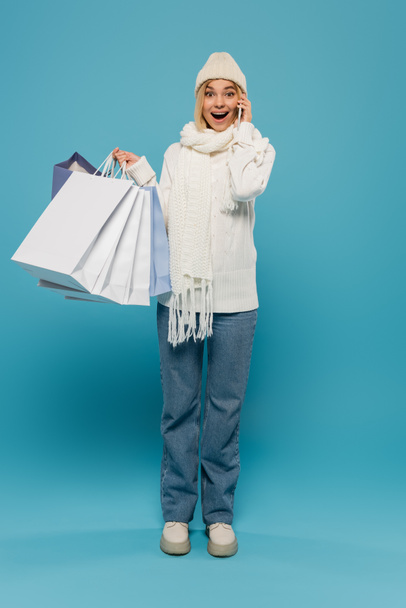 in voller Länge staunende Frau in weißem Pullover und Hut, die Einkaufstüten hält, während sie auf dem Smartphone auf blau spricht - Foto, Bild