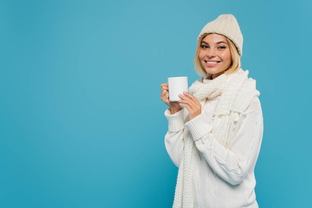 joyful young woman in white sweater and hat holding cup of coffee isolated on blue  - Φωτογραφία, εικόνα