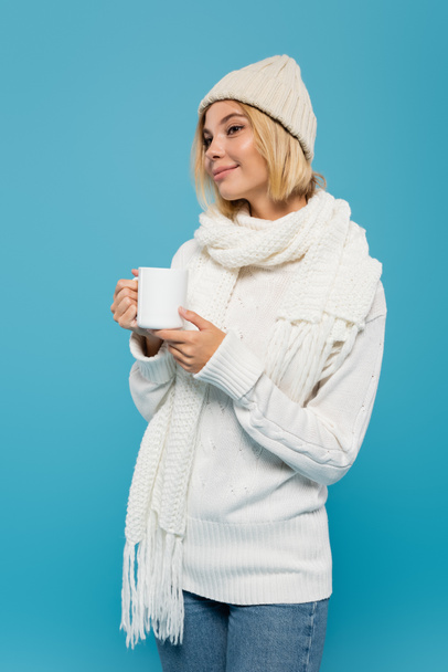 mujer sonriente en suéter blanco y sombrero de invierno sosteniendo taza de café aislado en azul  - Foto, Imagen