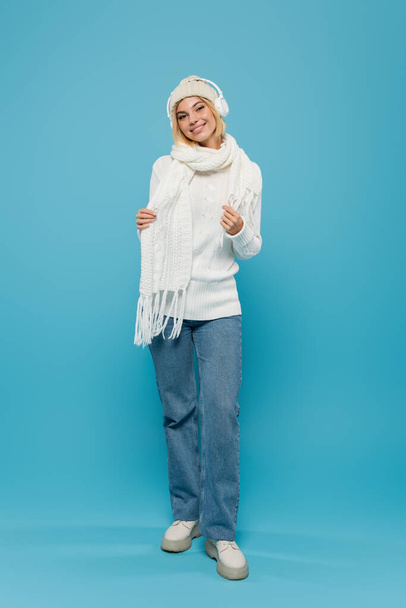 full length of smiling woman in white sweater and hat listening music in wireless headphones on blue  - Fotó, kép