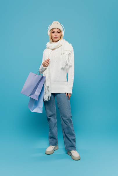 full length of young woman in winter outfit and wireless headphones holding shopping bags on blue  - Fotografie, Obrázek