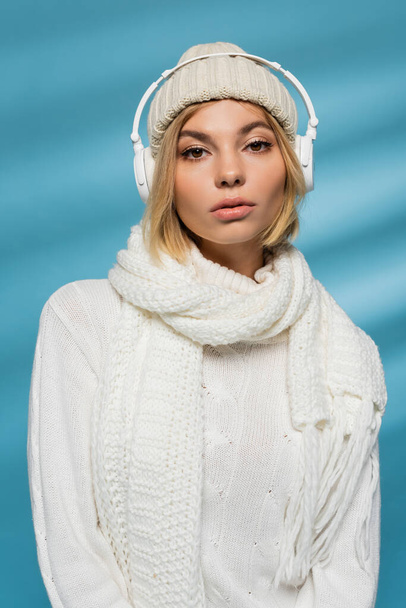 blonde woman in winter hat and wireless headphones listening music on blue  - Photo, Image