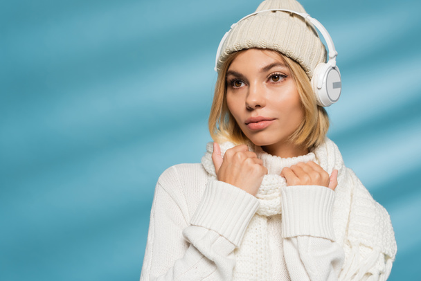 blonde young woman in knitted hat and wireless headphones looking away on blue  - Photo, Image