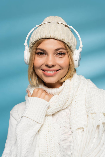 happy young woman in knitted hat and wireless headphones looking at camera on blue  - Photo, Image