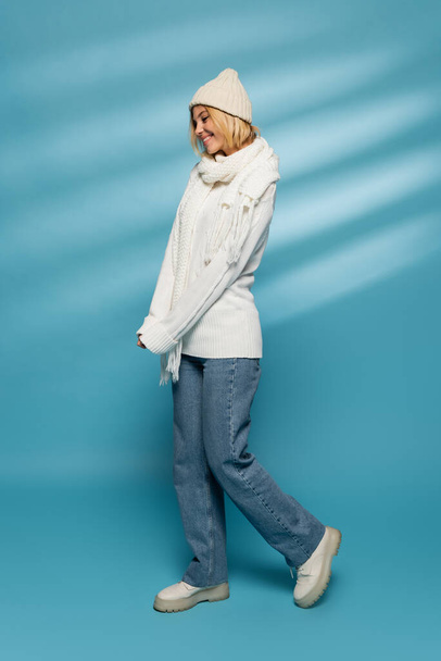 full length of happy young woman in knitted hat and jeans smiling on blue  - Fotó, kép