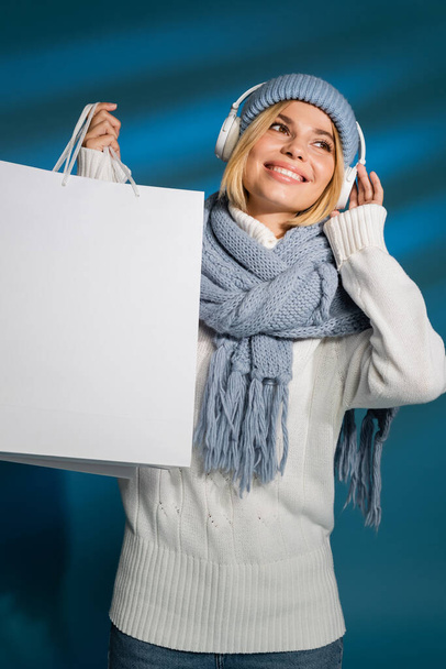 vidám fiatal nő télen sál és kalap hallgat zenét vezeték nélküli fejhallgató és gazdaság bevásárló táska kék  - Fotó, kép