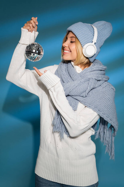 mujer alegre en sombrero de invierno y auriculares inalámbricos celebración de bola disco en azul  - Foto, Imagen