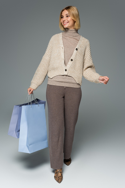 full length of cheerful blonde woman in turtleneck and cardigan holding shopping bags on grey - Photo, Image