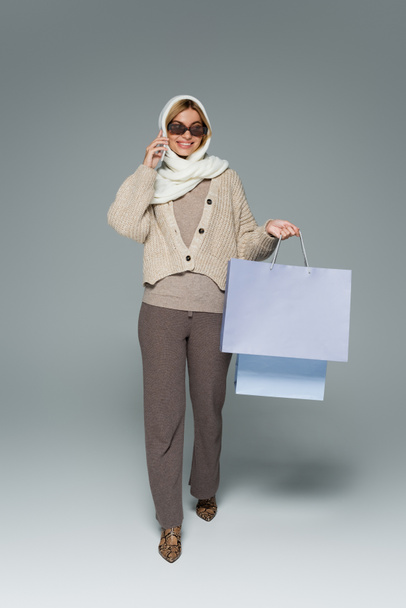 full length of cheerful woman in knitwear and sunglasses holding shopping bags while talking on smartphone on grey - Photo, Image