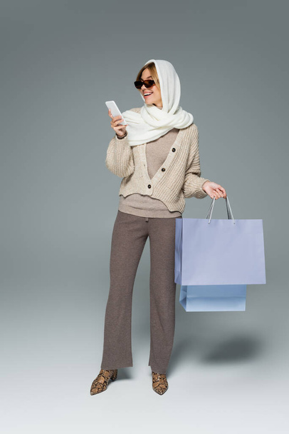 longitud completa de la mujer alegre en prendas de punto y gafas de sol sosteniendo bolsas de compras mientras se utiliza el teléfono inteligente en gris - Foto, Imagen