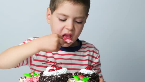 Beau garçon de 5 ans avec une cuillère mange une délicieuse crème d'un gâteau sur un fond blanc - Séquence, vidéo