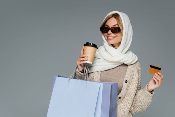 mujer feliz en gafas de sol sosteniendo taza de papel y tarjeta de crédito mientras está de pie con bolsas aisladas en gris - Foto, imagen