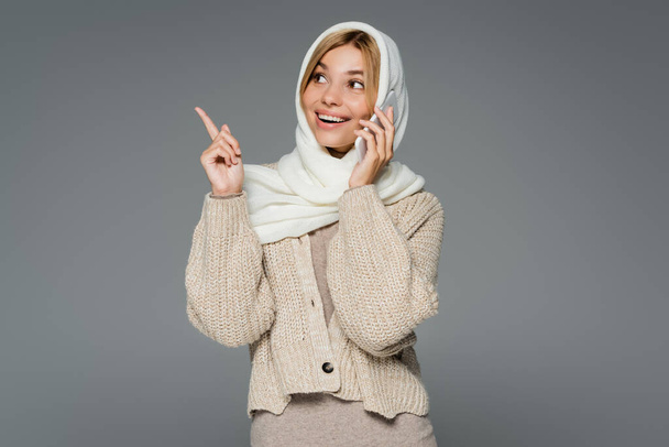 happy young woman in winter headscarf talking on mobile phone and pointing with finger isolated on grey - Foto, Imagem