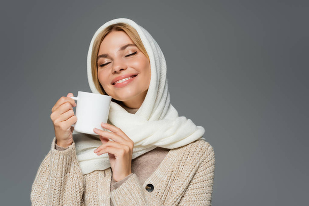 pleased woman in winter headscarf and knitted cardigan holding cup of coffee isolated on grey - Fotografie, Obrázek
