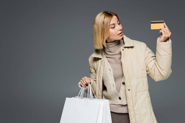 Young woman in warm outfit looking at credit card while holding shopping bags isolated on grey  - Φωτογραφία, εικόνα