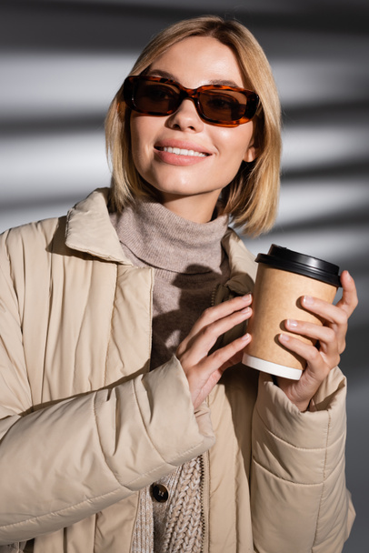Güneş gözlüklü ve kışlık ceketli gülümseyen kadın soyut gri arka planda kahve tutuyor.  - Fotoğraf, Görsel