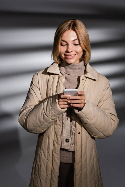 Pozytywna młoda kobieta w kurtce zimowej za pomocą telefonu komórkowego na abstrakcyjnym szarym tle  - Zdjęcie, obraz