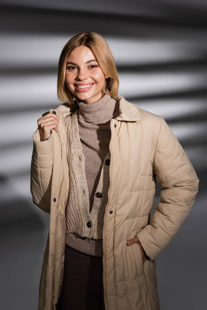 Χαρούμενη νεαρή γυναίκα με χειμερινό σακάκι κοιτάζοντας την κάμερα σε αφηρημένο γκρι φόντο  - Φωτογραφία, εικόνα