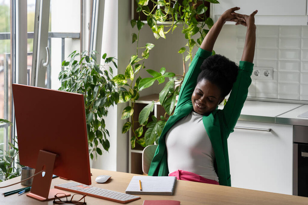 正のアフリカ系アメリカ人女性のオフィスワーカーがコンピュータとテーブルの上で働き、長い座り込み作業の後に暖まる。完成複合プロジェクトで喜び笑顔の若い女の子は自宅の植物と屋内にあります  - 写真・画像