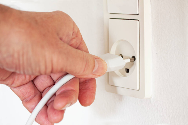 Close up of hand pulling cord out or in of modern white outlet on wall. Plug from an electrical appliance plugged into an electrical outlet on a white wall background. Electricity and power concept. - Φωτογραφία, εικόνα