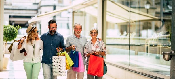 ομάδα τεσσάρων ανθρώπων που πηγαίνουν για ψώνια μαζί κρατώντας τσάντες ψώνια με δώρα ή δώρα για τα Χριστούγεννα - αγοράζοντας ρούχα και περισσότερο σε ένα εμπορικό κέντρο ή σε ορισμένα καταστήματα   - Φωτογραφία, εικόνα