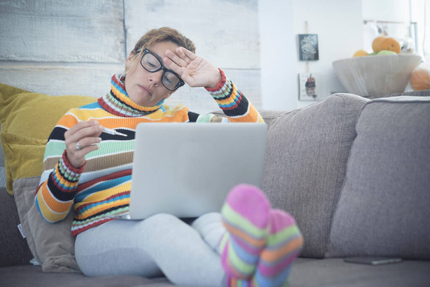 piękna kobieta siedząca w domu na kanapie - zmęczona kobieta z gorączką zdenerwowana serwetką w ręku - kobieta zmarznięta zimą - patrząca na termometr i z komputerem lub laptopem na nogach    - Zdjęcie, obraz