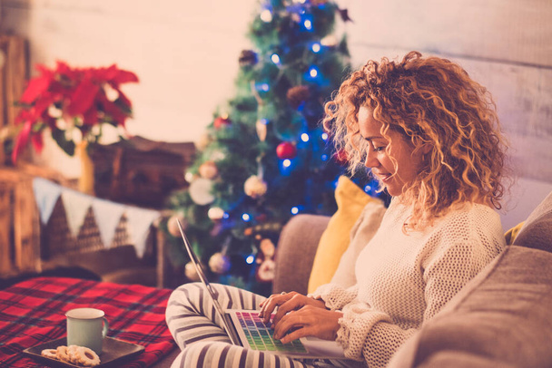 woman alone at home sitting on the sofa working or shopping with laptop and with cookies and tea or coffee and with Christmas tree at the background  - Photo, image