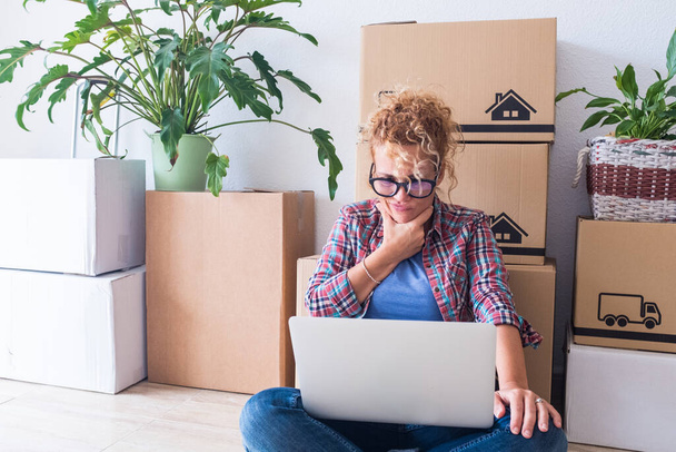 close up and portrait of young woman at home in a new house with boxes and packs in her back - adult woman using her laptop but not understanding what is in it  - Photo, Image