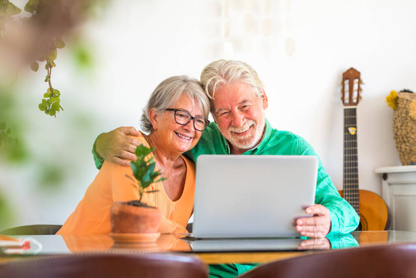 пара счастливых взрослых и пожилых людей или пожилых людей дома, сидя на диване наслаждаясь и весело вместе глядя и используя ноутбук или компьютер   - Фото, изображение
