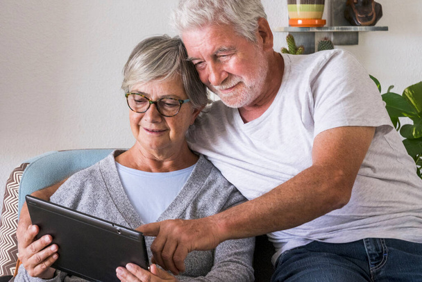 couple of seniors smiling and looking at the same tablet hugged on the sofa - indoor, at home concept - caucasians mature and retired man and woman using technology  - Foto, Imagem