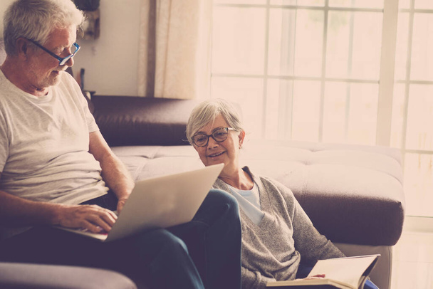 pareja de personas mayores en casa - hombre con portátil y gafas sentado en el sofá - mujer sentada en el suelo leyendo un libro - concepto de interior y amor  - Foto, imagen