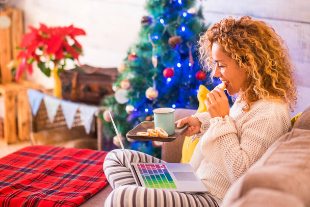 une belle femme bouclée annonce assis sur le canapé de travail ou en utilisant son ordinateur portable ou PC le jour de Noël à la maison célébrer - dame adulte manger des cookies et boire du thé  - Photo, image
