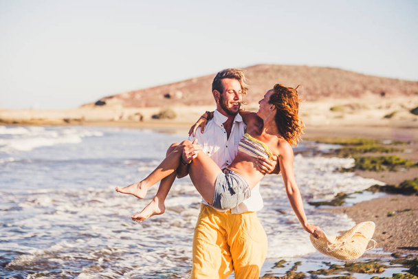 piękna para dwojga dorosłych na plaży spacerująca po piasku z kobietą na ramionach mężczyzny - mężczyzna trzymający swoją żonę na plaży  - Zdjęcie, obraz