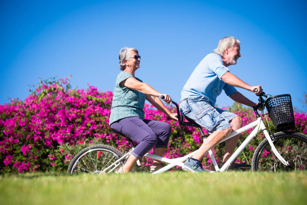 hermosa pareja de mayores y maduros casados, montando una bici doble, tándem, juntos para divertirse con un gran y soleado día - mujer y hombre   - Foto, Imagen