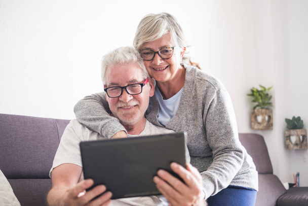 красивая пара пожилых людей или зрелых людей дома обнял глядя на тот же планшет или ноутбук - пенсионеры в очках, используя новую технологию   - Фото, изображение