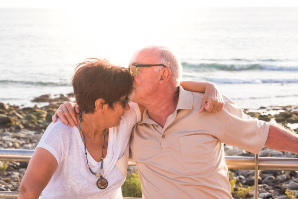 Seniorenpaar hockt mit viel Liebe am Strand - zusammen im Ruhestand - Frau mit Brille und Mann mit Meereshintergrund - küsst sie  - Foto, Bild