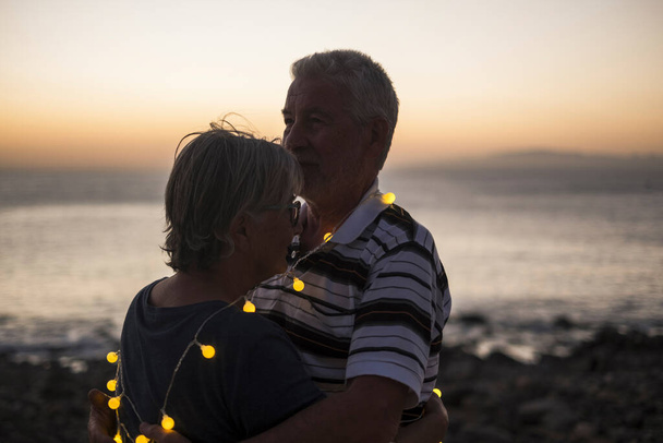 pareja de ancianos abrazándose en la playa con puesta de sol - pareja jubilada con luz alrededor de ellos - amor y momento de paz con puesta de sol  - Foto, imagen