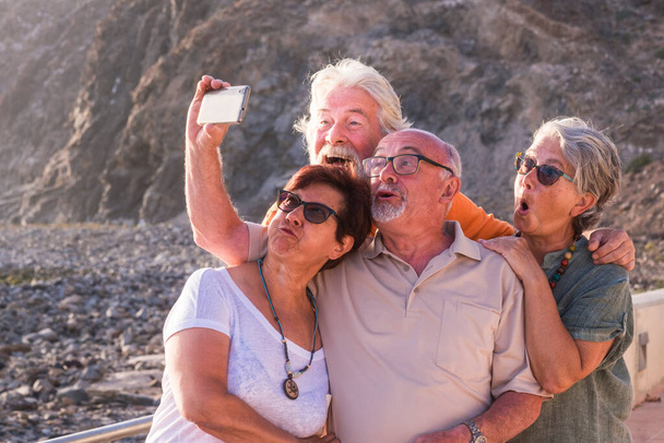 grupa czterech seniorów uśmiechających się i robiących głupie miny patrzących w obiektyw w selfie - przyjaźń dwóch par dojrzałych osób na plaży   - Zdjęcie, obraz