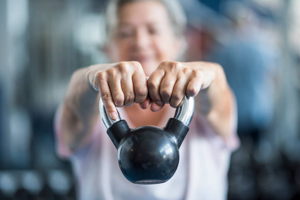primer plano de la persona mayor sosteniendo un peso con las manos delante de la cámara en el gimnasio - mujer adulta trabajando su cuerpo para estar saludable  - Foto, imagen