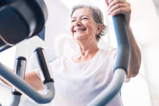 una mujer madura o mayor en el gimnasio de entrenamiento y hacer ejercicio en una máquina - activa pensionista estilo de vida y concepto  - Foto, Imagen