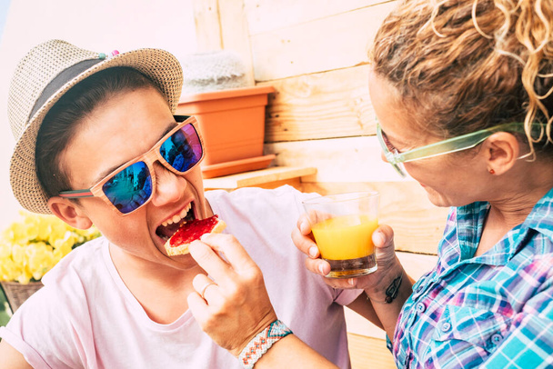 homme caucasien et femme adulte à la maison dans la terrasse prendre le petit déjeuner avec un drôle de visage de l'adolescent pendant qu'il mange un pain avec de la marmelade garçon avec des lunettes de soleil s'amuser et plaisanter  - Photo, image