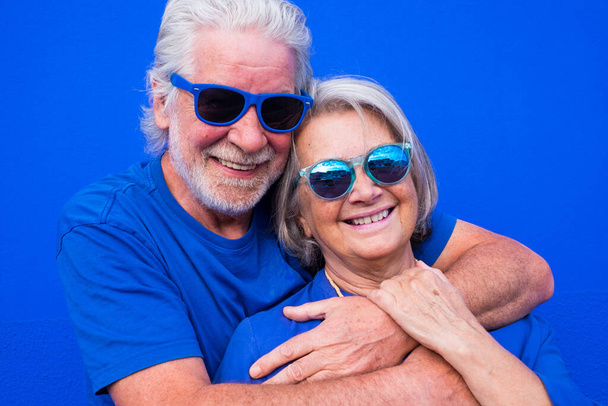Porträt zweier älterer Menschen oder Senioren, die gemeinsam Spaß haben, lächelnd in die Kamera schauen - Rentnerinnen und Rentner mit blauem Hemd und Sonnenbrille mit blauem Hintergrund - trendige, bunte und glückliche Menschen  - Foto, Bild