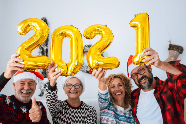 τέσσερα άτομα που διασκεδάζουν και χαμογελούν μαζί στο σπίτι κρατώντας ψηλά τους τέσσερις αριθμούς μπαλονιών του 2021 - οικογένεια γελώντας και γιορτάζοντας τα Χριστούγεννα και το νέο έτος   - Φωτογραφία, εικόνα