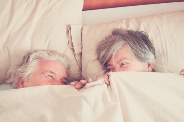 пара пожилых людей, глядя между и улыбаясь с любовью и влияют под одеялом на кровать - пенсионеры и зрелые взрослые женаты в спальне на утро, прежде чем встать  - Фото, изображение