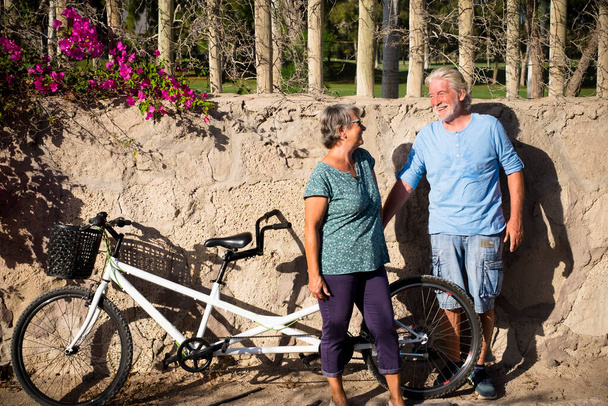 pareja de dos personas mayores y personas maduras de pie con la pared al fondo en un parque con su tándem o bici doble sonriendo y hablando  - Foto, imagen