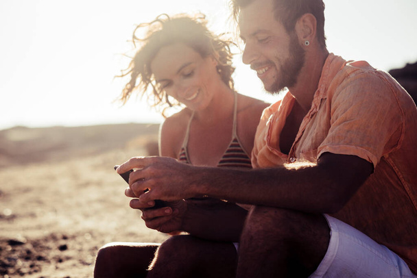 couple of adults at the beach talking and looking at the phone of the woman sitting on the rocks - woman in bikini looking at her phone and a man looking at the same phone at the sunset  - Photo, Image