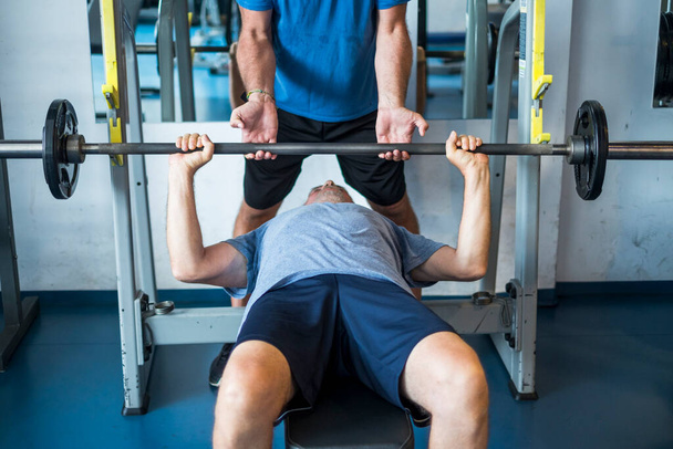 um homem maduro deitado no banco do ginásio treinando seu corpo para ser fitness e ativo sênior homem ajudando-o segurando o bar com as mãos  - Foto, Imagem