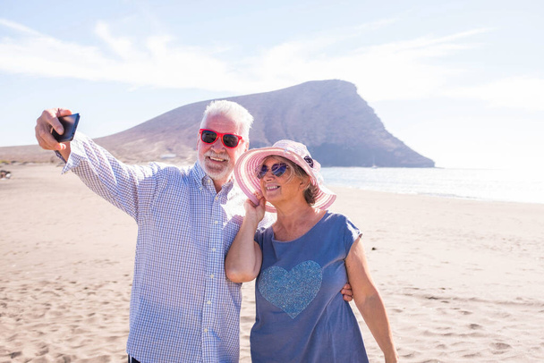 İki olgun insan sahilde selfie çekiyor gülümsüyor ve eğleniyor ve eğleniyor - yaşlılar ya da emekliler ya da tatillerinin tadını çıkarıyor - kumsalda güneşli bir gün.  - Fotoğraf, Görsel