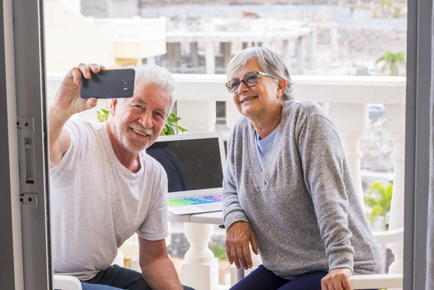 para seniorów na tarasie ich domu z laptopem i siedząc robiąc sobie selfie razem - zrelaksowana dojrzała emerytka i mężczyzna robiący zdjęcie  - Zdjęcie, obraz
