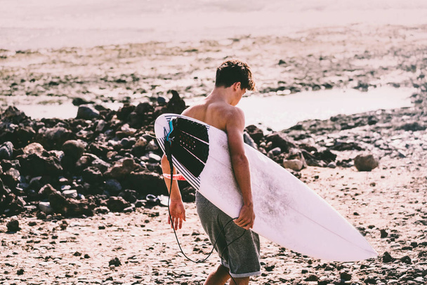 un adolescent entre à l'eau avec sa table de surf sous le bras prêt à aller surfer - athlète actif aller s'entraîner à l'eau  - Photo, image